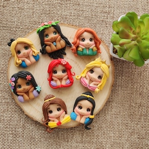 May include: Eight handmade polymer clay figurines of Disney princesses with different hairstyles and outfits. Each figurine has a different facial expression and is holding their hands together in front of their face. The figurines are arranged in a circle on a wooden surface.