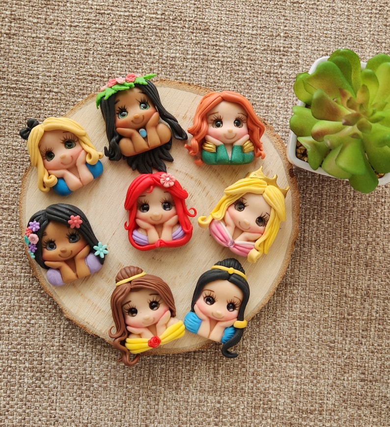 May include: Eight handmade polymer clay figurines of Disney princesses with different hairstyles and outfits. Each figurine has a different facial expression and is holding their hands together in front of their face. The figurines are arranged in a circle on a wooden surface.