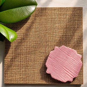 May include: A square coaster featuring a dark brown cross-hatched grid pattern on a tan base. A smaller, pink, scalloped-edge coaster with a textured, ribbed surface is placed on top of the larger square coaster, creating a layered visual effect.