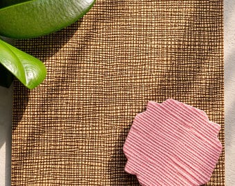 May include: A square coaster featuring a dark brown cross-hatched grid pattern on a tan base. A smaller, pink, scalloped-edge coaster with a textured, ribbed surface is placed on top of the larger square coaster, creating a layered visual effect.