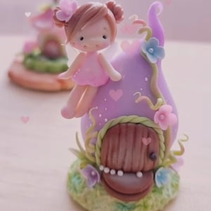 May include: A miniature fairy figurine with pink wings and a pink dress sits on top of a purple and green mushroom house with a brown door. The house is decorated with white dots and pink flowers. The background is a light pink with white hearts.