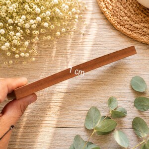 May include: A rectangular wooden block with a smooth, medium-brown finish. The side of the block is clearly marked with the text 1 cm, indicating its thickness. The object is held at an angle, showcasing its solid construction and flat edges.