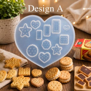 Puede incluir: Un molde de silicona translúcido en forma de corazón con varias formas hundidas para galletas, incluyendo estrellas, círculos, rectángulos y corazones. El texto "Design A" está impreso en blanco sobre el molde, rodeado de galletas horneadas.