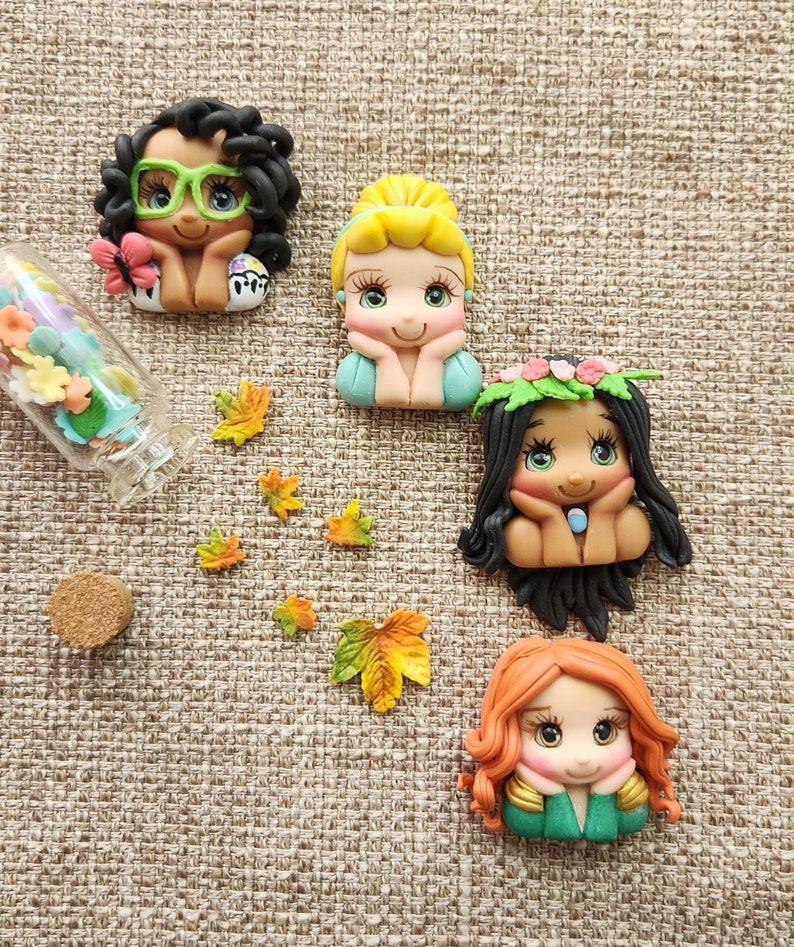 May include: Four handmade polymer clay figurines of cartoon characters with large eyes. The characters are all wearing different outfits and have different hair colors. The figurines are sitting on a brown burlap background.