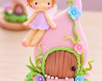 Könnte beinhalten: Eine Tonfigur mit einer kleinen Fee mit braunen Haaren in Dutts, die ein hellviolettes Kleid trägt und auf einem rosa tropfenförmigen Haus mit brauner Tür, grünen Ranken sowie blauen und rosa Blumen auf einem grünen Sockel sitzt.