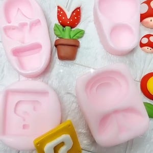 以下が含まれることがあります： ピンク色のシリコンモールドとフォンダンデコレーションのコレクション。モールドには、靴、花、疑問符の形が含まれています。フォンダンデコレーションには、赤と白の水玉模様の花、茶色のポット、キノコと疑問符のブロックが含まれています。