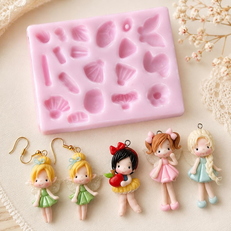 Puede incluir: Un molde de silicona rosa rectangular con varias formas peque&ntilde;as en relieve para manualidades, acompa&ntilde;ado de cinco dijes en miniatura con tem&aacute;tica de hadas, incluyendo dos pendientes con figuras de pelo amarillo y tres colgantes de personajes.