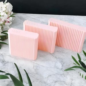 Puede incluir: Tres jaboneras rectangulares de color rosa claro con ranuras verticales. Las jaboneras están dispuestas sobre una superficie de mármol, con flores blancas y hojas verdes en el fondo. El texto "NMM" es visible en la parte superior de cada jabonera.
