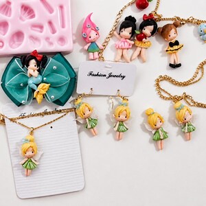 Puede incluir: Una colecci&oacute;n de joyer&iacute;a con figuras en miniatura de hadas y personajes, que incluye collares dorados, pendientes en una tarjeta de Fashion Jewelry, un lazo para el pelo color verde azulado con una sirena y un molde de silicona rosa con formas.