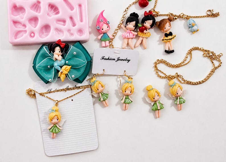 Puede incluir: Una colecci&oacute;n de joyer&iacute;a con figuras en miniatura de hadas y personajes, que incluye collares dorados, pendientes en una tarjeta de Fashion Jewelry, un lazo para el pelo color verde azulado con una sirena y un molde de silicona rosa con formas.