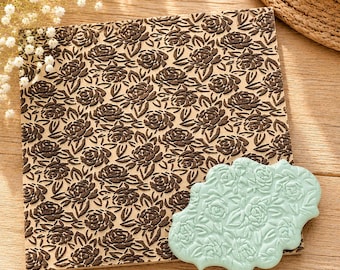 May include: A square wooden embossing plate featuring a repeating dark brown rose and leaf pattern, paired with a smaller, light mint green scalloped plaque that displays the same intricate floral design in a raised, embossed texture.