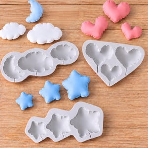 May include: Three grey silicone moulds for crafting shapes, including a cloud and moon mould, a heart mould, and a star mould. Accompanying the moulds are finished pieces: a light blue moon, two white clouds, three pink hearts, and three light blue stars.