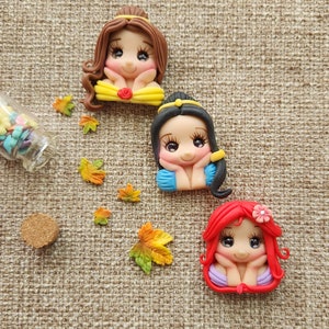 May include: Three handmade polymer clay figurines of Disney princesses. Each figurine is a different princess with a different hair color and dress. The princesses are all smiling and have their hands on their chins. The figurines are on a brown burlap background.