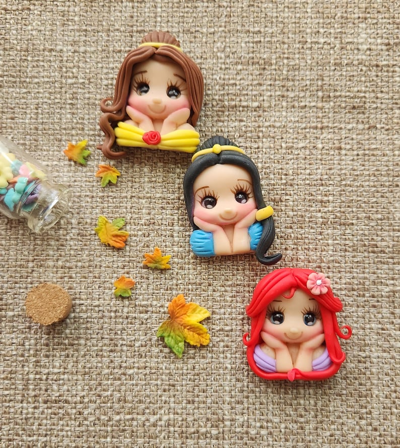 May include: Three handmade polymer clay figurines of Disney princesses. Each figurine is a different princess with a different hair color and dress. The princesses are all smiling and have their hands on their chins. The figurines are on a brown burlap background.