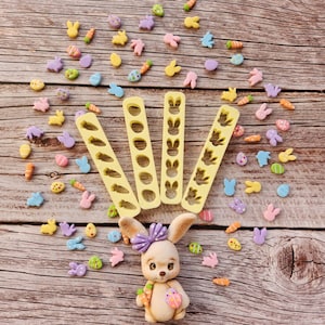 May include: A set of five yellow plastic cookie cutters with different Easter-themed designs, including bunnies, eggs, and carrots. The cutters are arranged in a fan shape on a wooden surface. There are also small, colourful fondant decorations scattered around the cutters, including bunnies, eggs, and carrots.