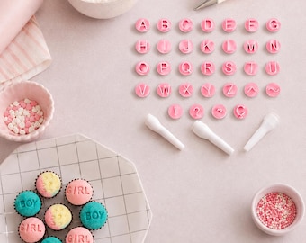 Könnte beinhalten: Ein Set aus rosa runden Alphabetstempeln mit den Buchstaben A bis Z, Satzzeichen und drei weißen Kunststoffgriffen. Ebenfalls enthalten sind kleine Cupcakes mit blauem und rosa Zuckerguss, beschriftet mit boy oder girl, sowie Streusel.