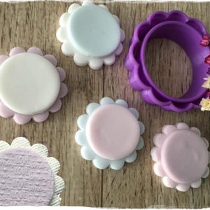 May include: A collection of pastel-colored, flower-shaped fondant decorations and a purple cookie cutter. The decorations include white, light blue, and pink circles with scalloped edges. One decoration has the word "LOVE" embossed on it.