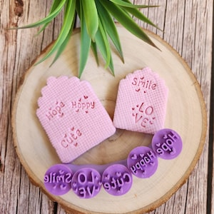 May include: A set of pink and purple rubber stamps with various words and designs, including "Hope", "Happy", "Cute", "Love", "Smile", and hearts. The stamps are arranged on a wooden surface.