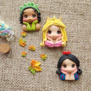 May include: Three handmade polymer clay figurines of Disney princesses: Tiana, Snow White, and Aurora. Each figurine has a detailed face with painted eyes and a sweet expression. The figurines are set against a brown burlap background.