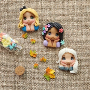 May include: Three handmade polymer clay dolls with painted faces and floral accents. The dolls are wearing blue, purple, and white dresses. The dolls are sitting on a brown fabric background.