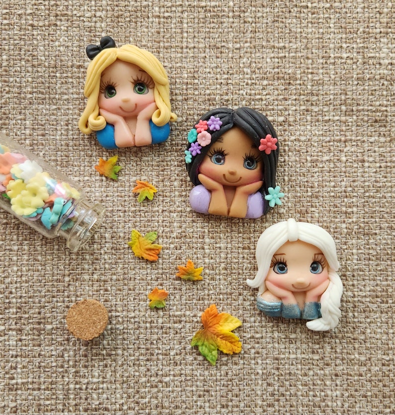 May include: Three handmade polymer clay dolls with painted faces and floral accents. The dolls are wearing blue, purple, and white dresses. The dolls are sitting on a brown fabric background.
