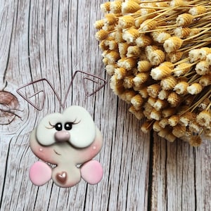 Puede incluir: Figurita de arcilla hecha a mano de un insecto estilizado en rosa y blanco. El insecto tiene ojos grandes blancos, orejas rosas y un detalle en forma de corazón. Un ramo de flores secas se encuentra en el fondo.