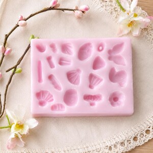 Puede incluir: Un molde de silicona rectangular de color rosa claro que presenta varias formas empotradas, incluyendo peque&ntilde;os p&eacute;talos de flores, hojas y siluetas de mariposas. El molde est&aacute; dise&ntilde;ado para manualidades y se presenta como una l&aacute;mina plana y flexible con m&uacute;ltiples cavidades.
