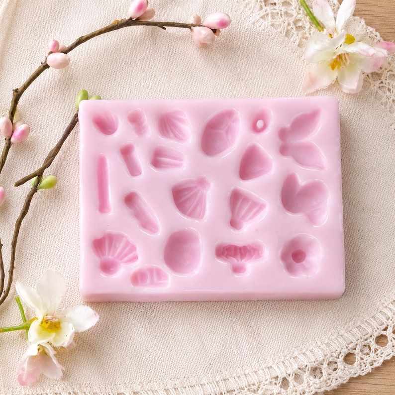 Puede incluir: Un molde de silicona rectangular de color rosa claro que presenta varias formas empotradas, incluyendo peque&ntilde;os p&eacute;talos de flores, hojas y siluetas de mariposas. El molde est&aacute; dise&ntilde;ado para manualidades y se presenta como una l&aacute;mina plana y flexible con m&uacute;ltiples cavidades.