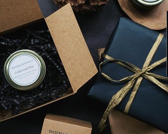 Puede incluir: Vista cenital de velas perfumadas y cajas de regalo. Una lata de vela con "Cedarwood Pine" está en una caja abierta. Un regalo envuelto está atado con una cinta dorada. La escena está sobre una superficie oscura.