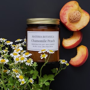 May include: A brown glass candle jar with a gold lid. The label reads "MATERIA BOTANICA Chamomile Peach HANDCRAFTED SOY WAX CANDLE Net Wt. 7.5 oz. Burn Time: 50 hrs.". The candle is surrounded by white daisy flowers and peach slices on a black background.