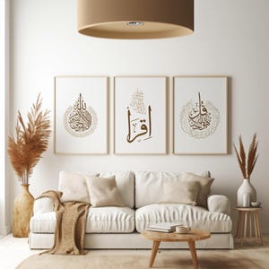 May include: Three framed art prints with Arabic calligraphy in brown tones hang above a white sofa. The room features a neutral color palette with a beige rug, wooden coffee table, and a large tan drum pendant light.