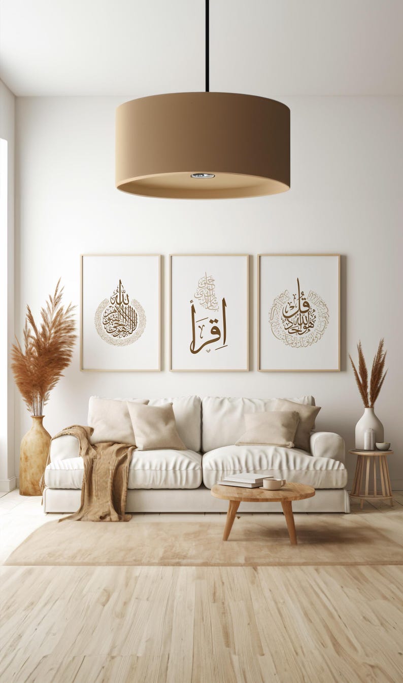May include: Three framed art prints with Arabic calligraphy in brown tones hang above a white sofa. The room features a neutral color palette with a beige rug, wooden coffee table, and a large tan drum pendant light.