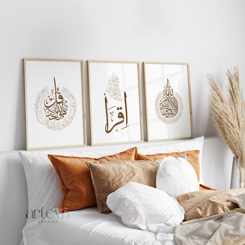 May include: Three framed art prints with Arabic calligraphy in brown tones. The prints are displayed above a bed with orange and tan pillows and white bedding. The frames are light-colored wood.