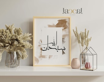 May include: A framed art print with Arabic calligraphy in black script on a white background, set against a beige and brown abstract design. The artwork is displayed on a white shelf with decorative elements, including dried flowers in a glass vase and a geometric metal house with a pink vase inside.