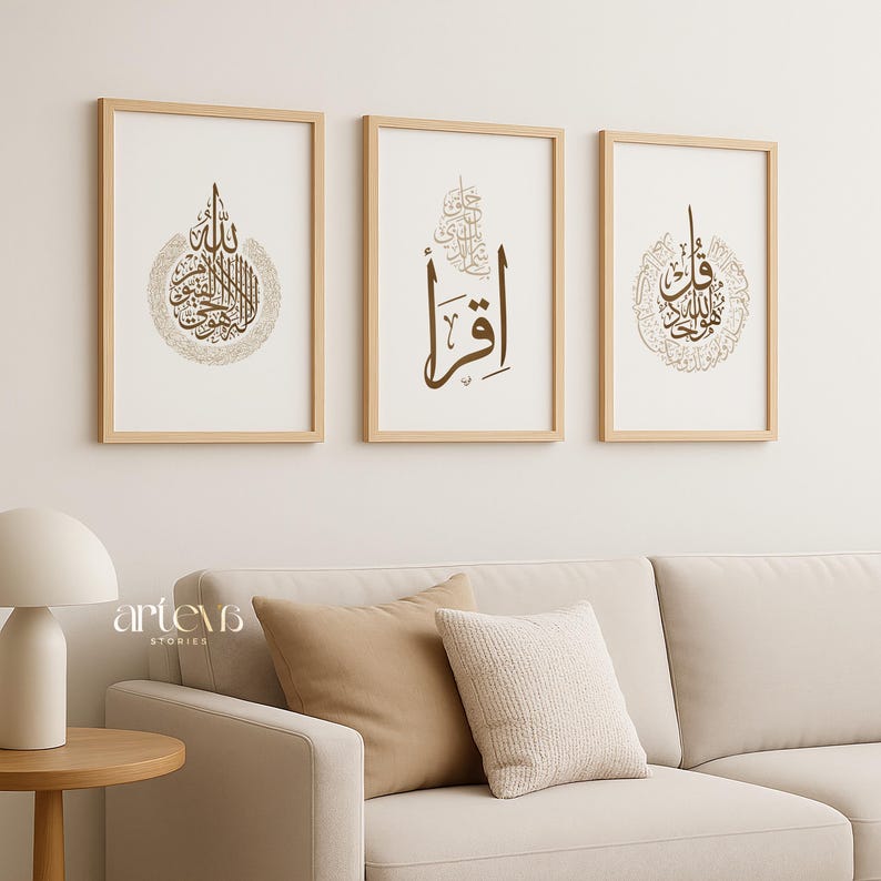 May include: Three framed art prints with Arabic calligraphy in shades of brown and gold. The prints are displayed above a beige sofa with decorative pillows. The frames are made of light-colored wood, and the overall aesthetic is minimalist and elegant.