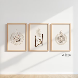 May include: Three framed art prints with Arabic calligraphy in brown tones. The prints are displayed on a white wall with a wooden floor. The frames are made of light wood. The calligraphy is in a decorative style.