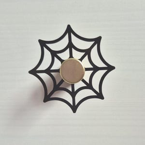 以下が含まれることがあります： 黒いクモの巣のキャビネットノブ、金色の中心部。装飾的なノブは家具のユニークなアクセントで、ハロウィンやゴシック様式を添えます。白い表面に取り付けられています。