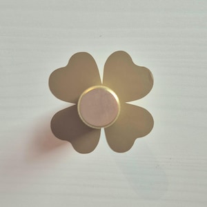 May include: A four-leaf clover-shaped cabinet knob. The petals are a muted gold colour, with a light pink circle at the centre, framed by a gold ring. This decorative knob is designed for use on cabinets or drawers, adding a touch of elegance.