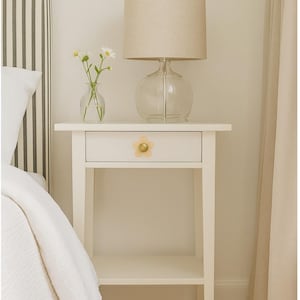 May include: A cream-coloured bedside table with a drawer and a shelf. A clear glass lamp with a beige shade is on top. A small vase with white flowers sits beside the lamp. The drawer has a flower-shaped knob.