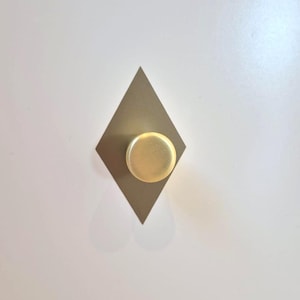 May include: A gold-colored, round cabinet knob is centered on a diamond-shaped, muted gold plate. The knob and plate are mounted on a white surface, creating a minimalist design.