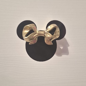 May include: A gold-coloured bow-shaped decorative element is centered above two black circles, resembling a famous cartoon character. The bow has a detailed, textured surface. The background is a white, wood-grain pattern.