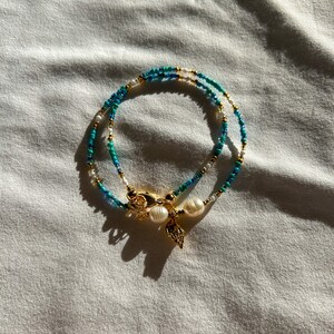 May include: A layered necklace with blue and white beads and a gold charm with two white pearls. The necklace is on a white fabric background.