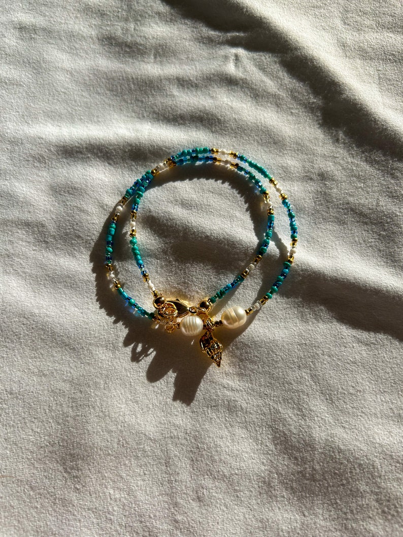 May include: A layered necklace with blue and white beads and a gold charm with two white pearls. The necklace is on a white fabric background.