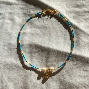 May include: A gold necklace with blue and white beads and a gold seashell charm. The necklace is on a white fabric background.