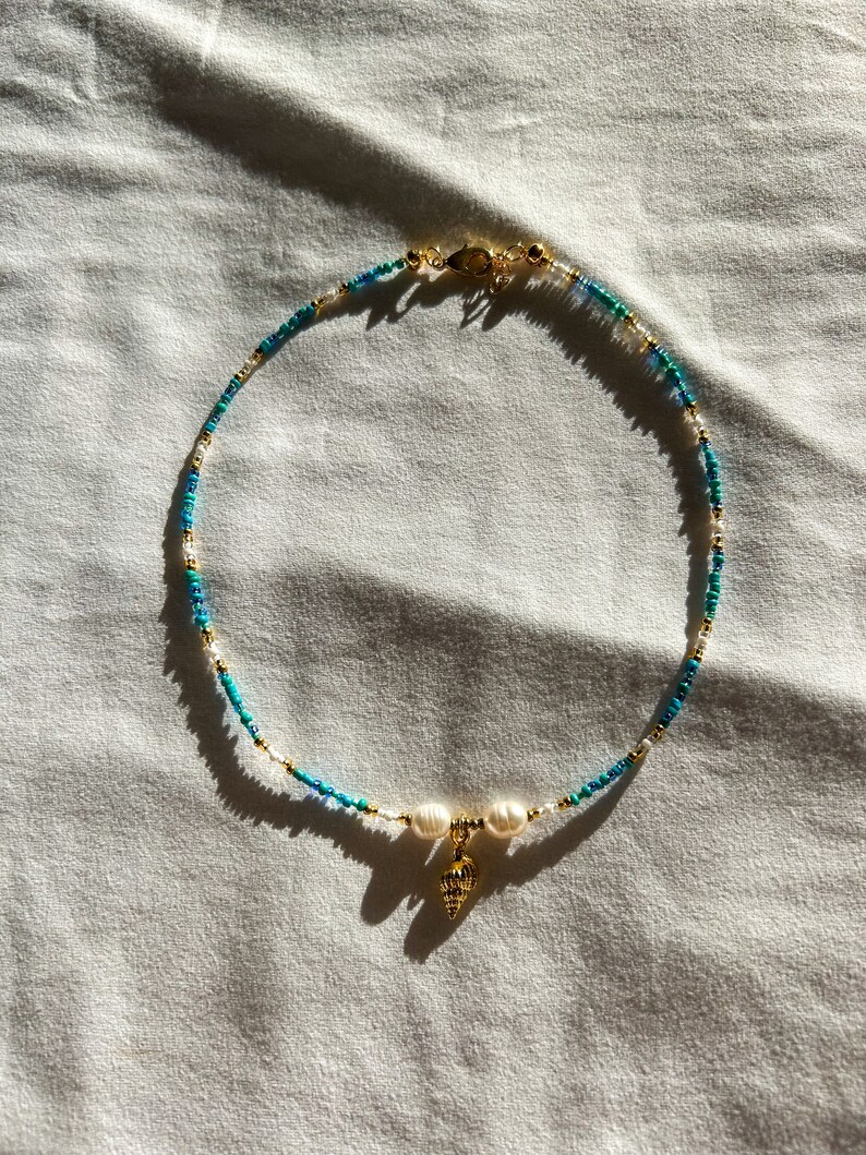 May include: A gold necklace with blue and white beads and a gold seashell charm. The necklace is on a white fabric background.