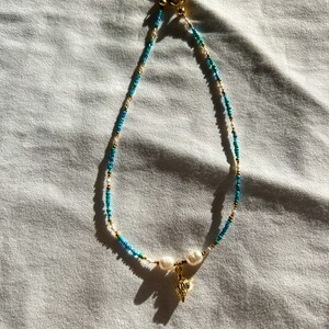 May include: A delicate gold chain necklace with white and blue beads and a small gold seashell charm. The necklace is on a white fabric background.