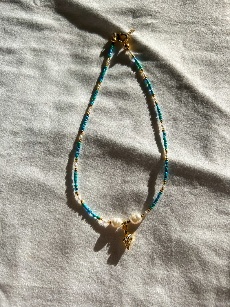 May include: A delicate gold chain necklace with white and blue beads and a small gold seashell charm. The necklace is on a white fabric background.
