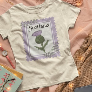 May include: A cream-coloured t-shirt with a postage stamp design featuring a thistle illustration. The stamp reads "Scotland" and "First Class". The design includes a purple border and a green and purple thistle.