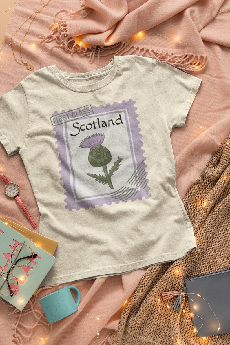 May include: A cream-coloured t-shirt with a postage stamp design featuring a thistle illustration. The stamp reads "Scotland" and "First Class". The design includes a purple border and a green and purple thistle.