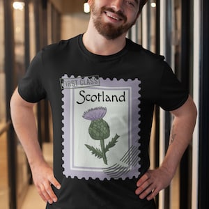 May include: Black t-shirt with a postage stamp design. The stamp shows a thistle and the words "First Class" and "Scotland". The thistle is purple and green. The t-shirt is worn by a person.